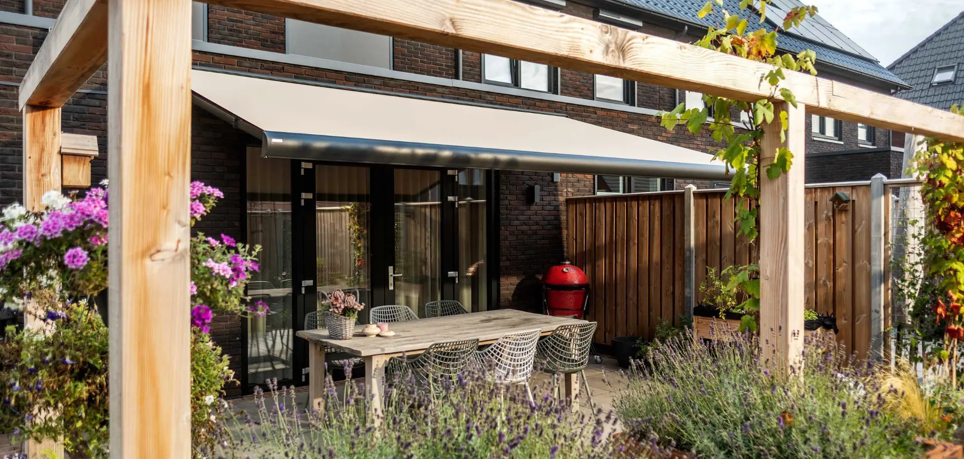 Beige uitvalscherm boven een terras met eettafel, tussen een houten pergola