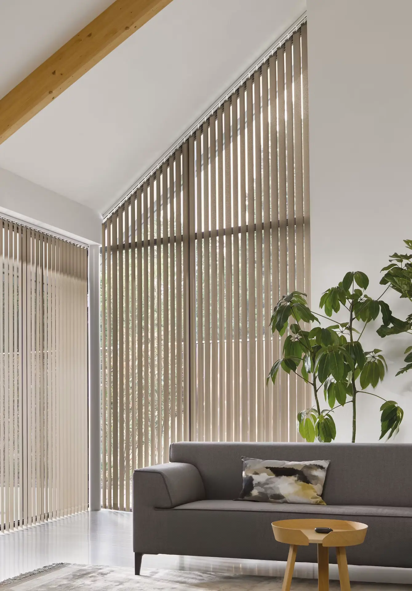 Verticale lamellen in beige stof op een schuin, hoog raam in een lichte woonkamer met grijze bank en groene plant