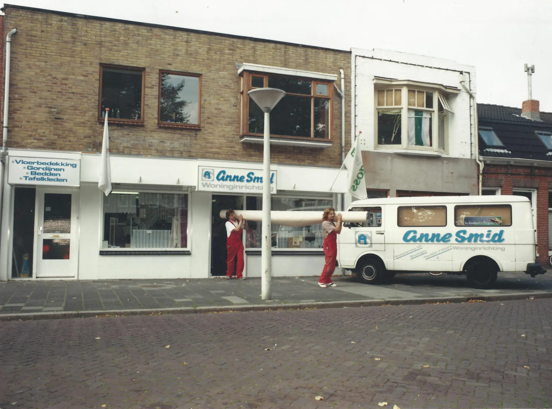 Oude foto van de winkel van Anne Smid, met twee medewerkers in rode overalls die een rol vloerbedekking dragen, naast de bestelbus